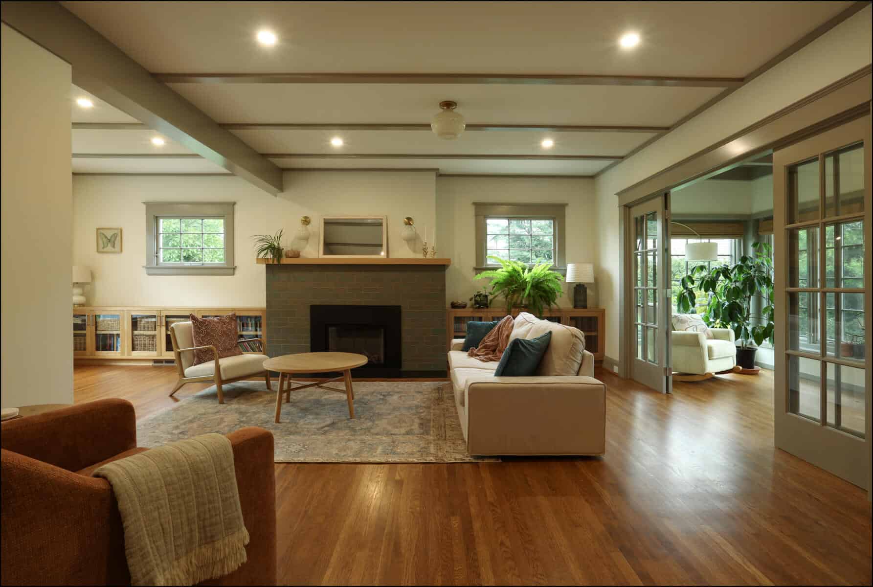 A spacious living room with wooden floors, a brick fireplace, a beige sofa, an orange armchair, and large windows. Bookshelves and green plants decorate the space. Spacious living room featuring a modern fireplace, comfortable seating, and natural light, showcasing ReCraft Home Remodeling's whole house remodel in Portland, Oregon.