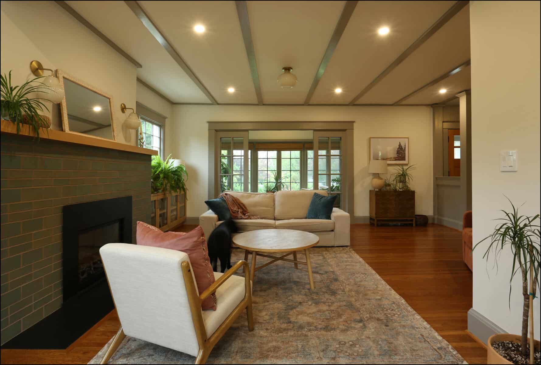 A cozy living room with a fireplace, beige armchair, white sofa, round wooden coffee table, and potted plants. Warm lighting and wooden flooring complete the inviting atmosphere. Cozy living room featuring a white sofa with colorful pillows, a round coffee table, a fireplace with green tiles, and large windows allowing natural light, showcasing a whole house remodel by ReCraft Home Remodeling in Portland, Oregon.