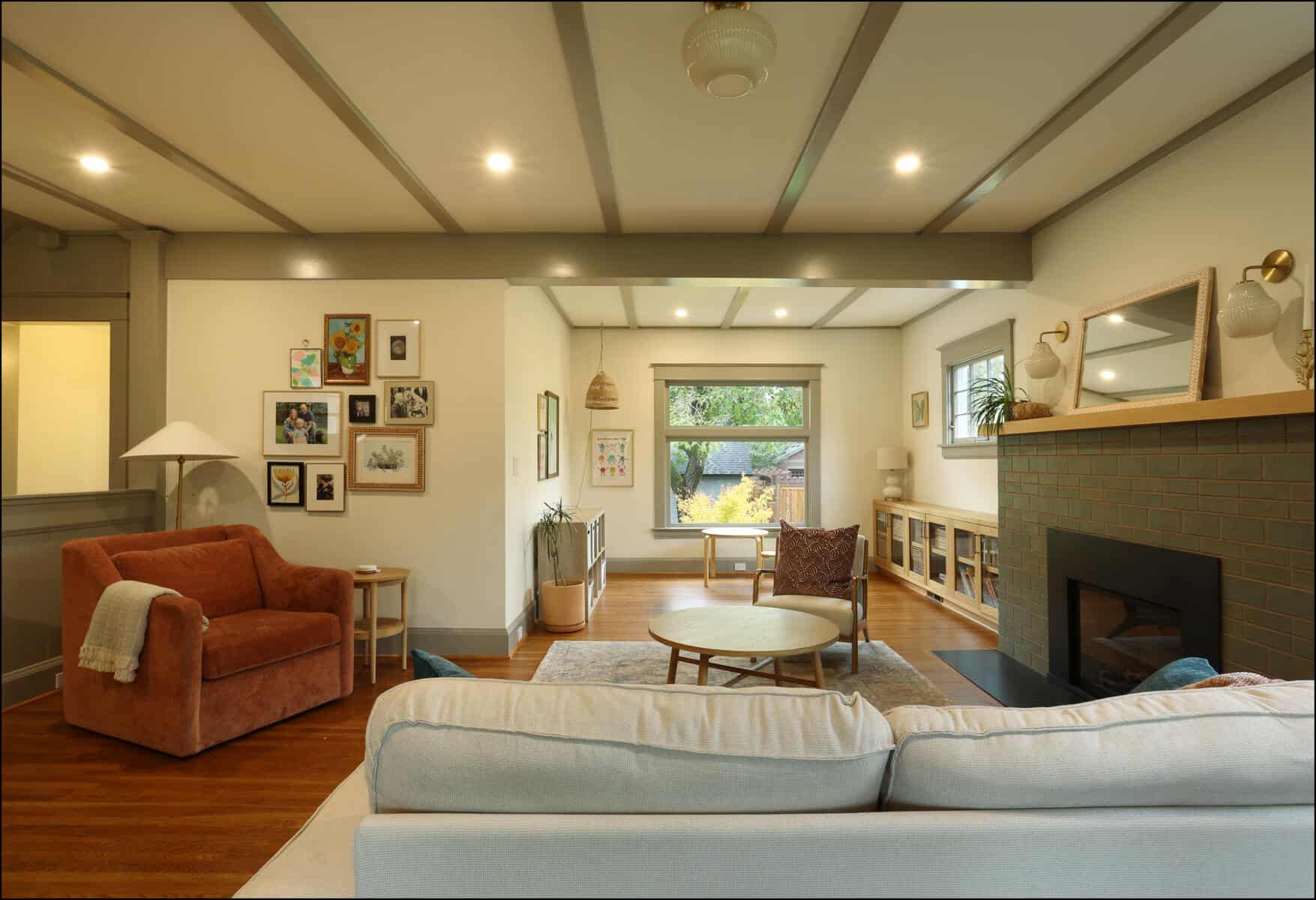 A cozy living room with a beige sofa, orange armchair, round wooden coffee table, and a fireplace. Artwork decorates the walls, and large windows offer a view of greenery outside. Living room with white sectional sofa, red armchair, wooden flooring, and framed artwork on walls, showcasing a whole house remodel by ReCraft Home Remodeling in Portland, Oregon.