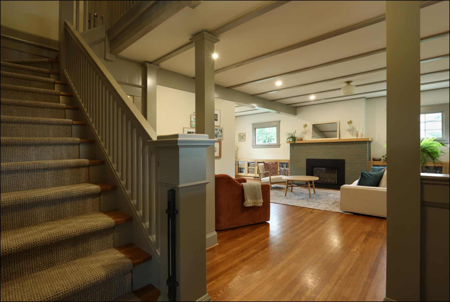 A cozy living room with a fireplace, sofa, and armchair. Wooden floors and a staircase are visible. The room has soft lighting and framed artwork on the walls. Whole house remodel showcasing an open living area with hardwood flooring, modern furnishings, and a staircase, emphasizing ReCraft Home Remodeling's design expertise in Portland, Oregon.