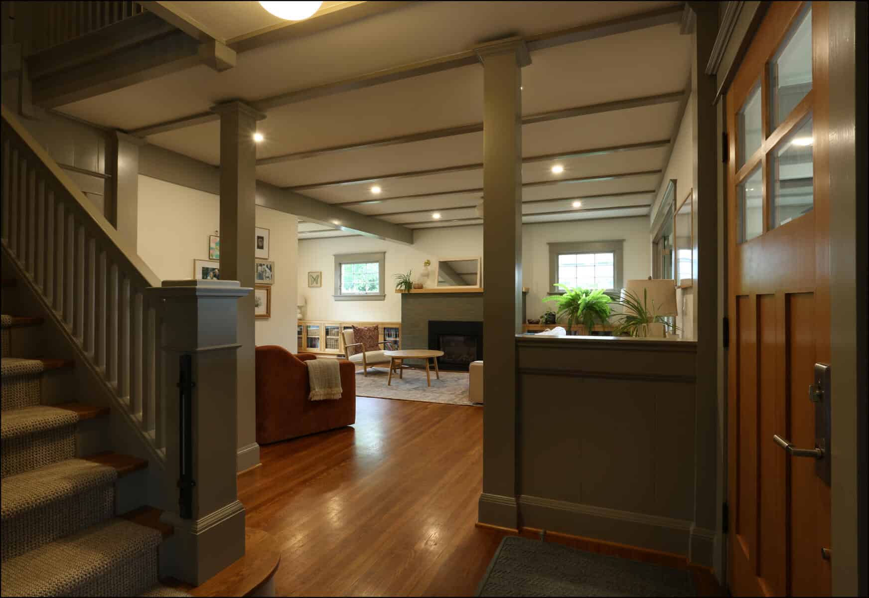 A cozy living room with wooden floors, a staircase on the left, a fireplace, and comfortable seating. Walls are painted in neutral tones with soft lighting illuminating the space. Welcoming entryway leading to an open living space with wooden floors, modern furnishings, and natural light, showcasing a whole house remodel by ReCraft Home Remodeling in Portland, Oregon.