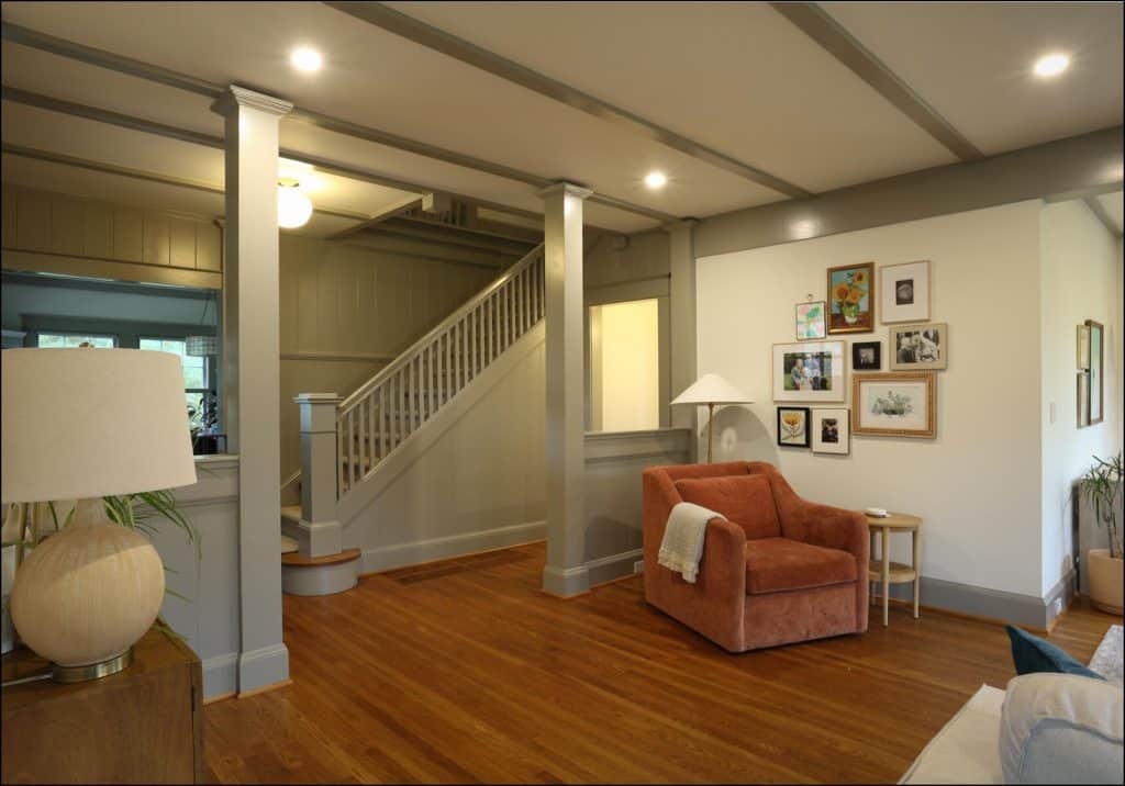 A cozy living room with an orange armchair, a side table, and framed pictures on the wall. Wooden flooring and gray-painted walls and ceiling beams. Stairs are visible in the background. A cozy living room with an orange armchair, a side table, and framed pictures on the wall. Wooden flooring and gray-painted walls and ceiling beams. Stairs are visible in the background.