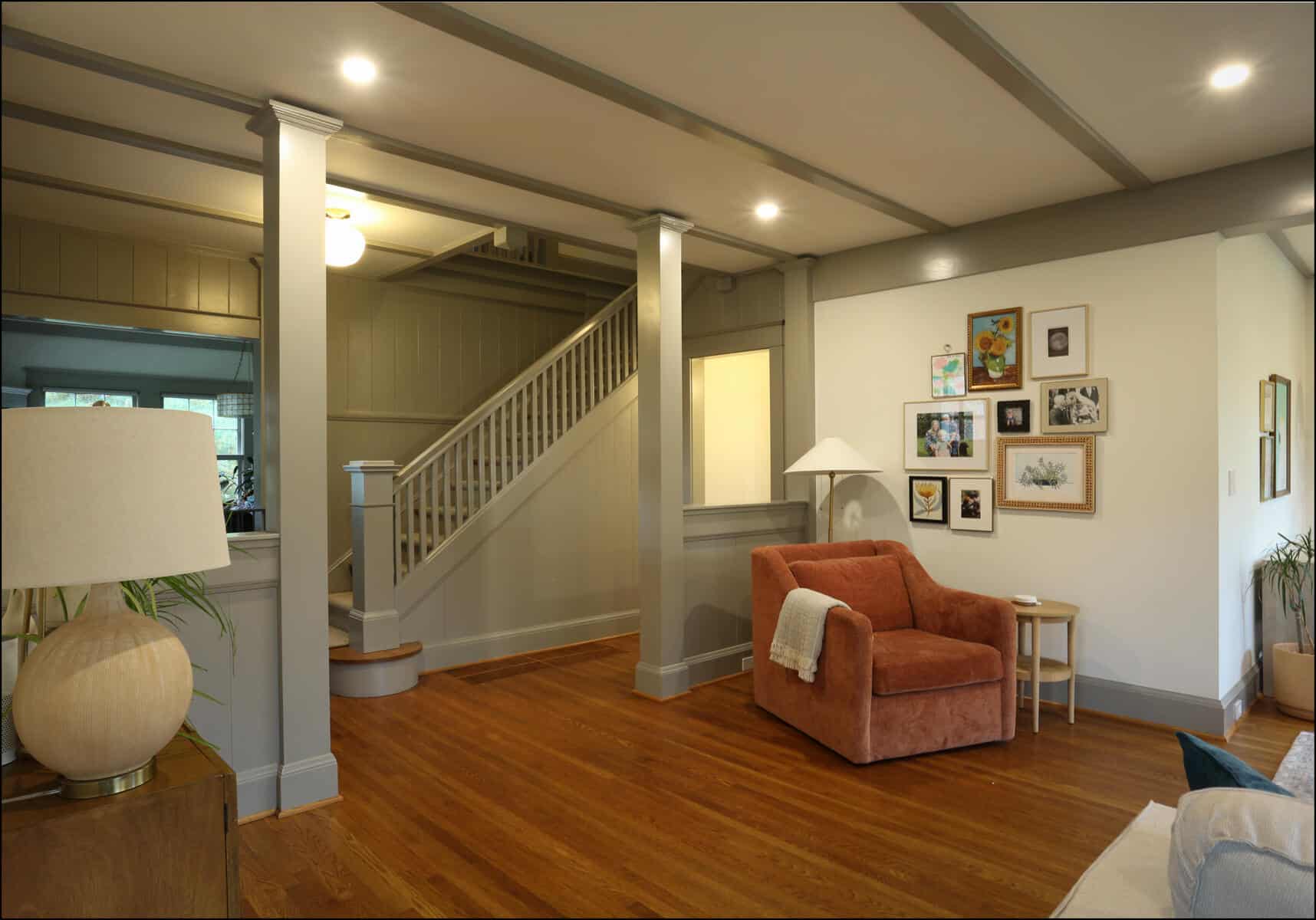 A cozy living room with an orange armchair, a side table, and framed pictures on the wall. Wooden flooring and gray-painted walls and ceiling beams. Stairs are visible in the background. Cozy living room featuring a pink armchair, decorative wall art, and a staircase, showcasing ReCraft Home Remodeling's whole house remodeling project in Portland, Oregon.