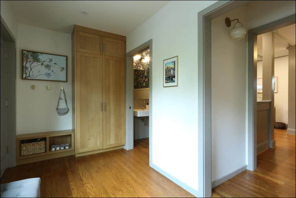 A hallway with wooden floors, a closet, a framed painting, a wall-mounted bag, shelves with shoes, and a view into a bathroom. A hallway with wooden floors, a closet, a framed painting, a wall-mounted bag, shelves with shoes, and a view into a bathroom.
