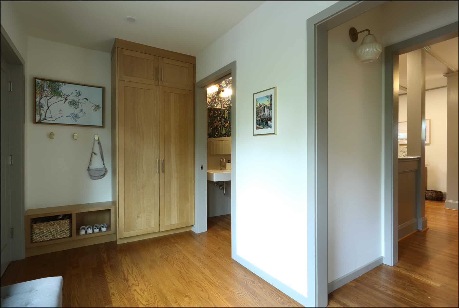 A hallway with wooden floors, a closet, a framed painting, a wall-mounted bag, shelves with shoes, and a view into a bathroom. Interior view of a modern hallway featuring wooden cabinetry, framed artwork, and an open doorway leading to a bathroom, highlighting ReCraft Home Remodeling's design aesthetic in whole house renovations.