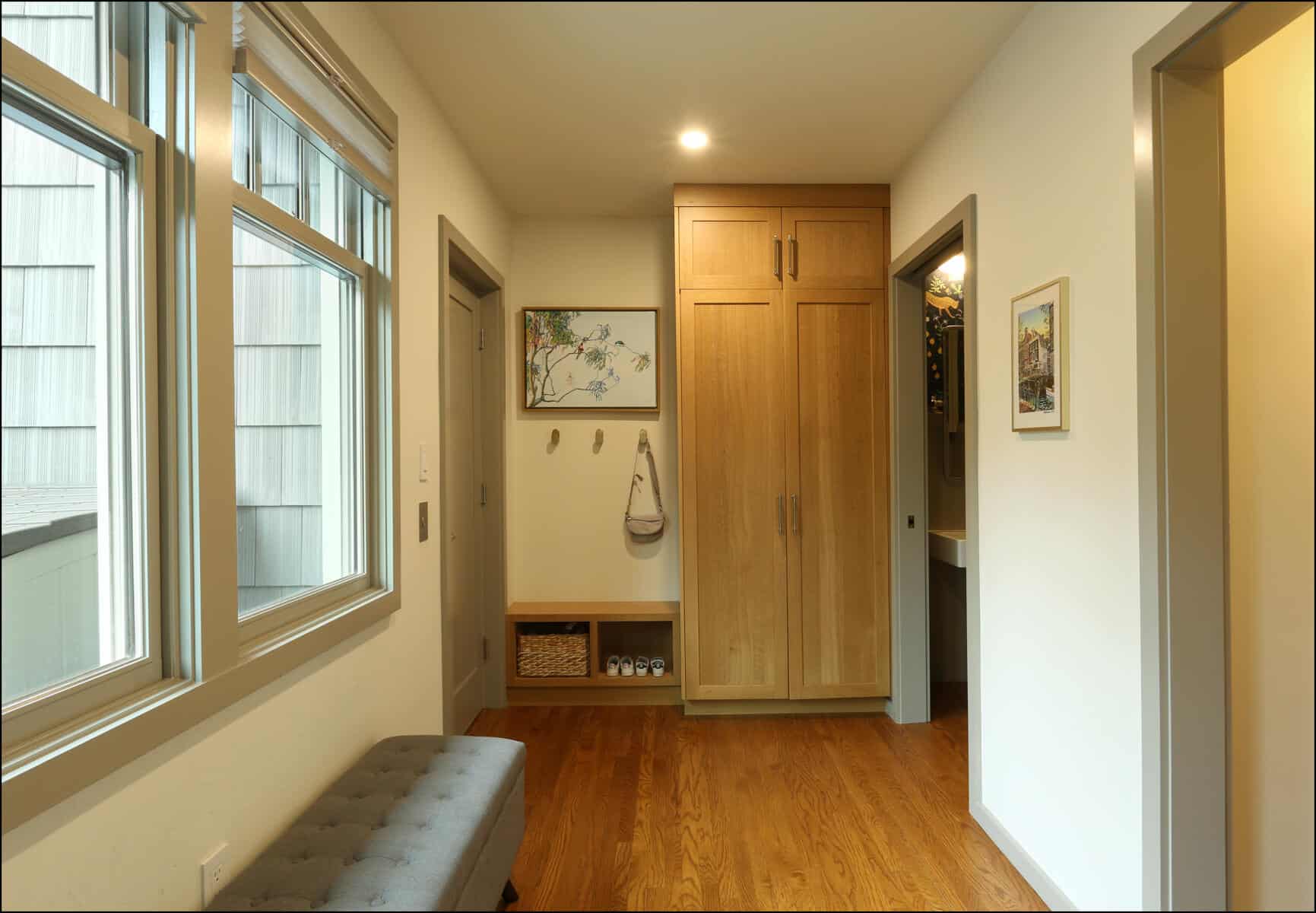 A hallway with a wooden floor, a bench, a wardrobe, coat hooks, a small basket shelf, a framed picture, and a window. Modern hallway featuring wooden flooring, built-in storage, and natural light from large windows, showcasing ReCraft Home Remodeling's whole house remodel in Portland.