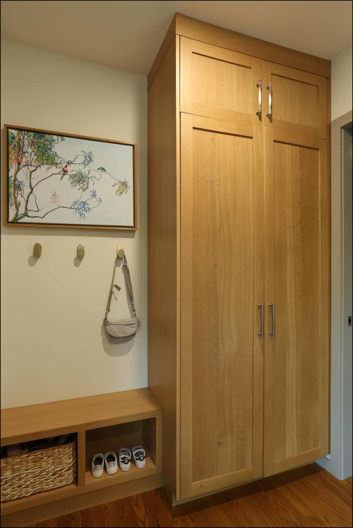 A wooden storage unit with cabinets and shelves in a hallway. A painting, hooks, a hanging bag, and shoes are arranged nearby. Modern entryway with wooden cabinetry, shoe storage, and wall hooks, showcasing ReCraft Home Remodeling's design expertise in whole house remodels in Portland, Oregon.