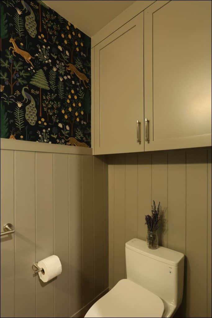 A small bathroom with taupe paneling, a white toilet, floral and animal-patterned wallpaper, cabinetry above, and a glass jar with lavender on the tank. A small bathroom with taupe paneling, a white toilet, floral and animal-patterned wallpaper, cabinetry above, and a glass jar with lavender on the tank.