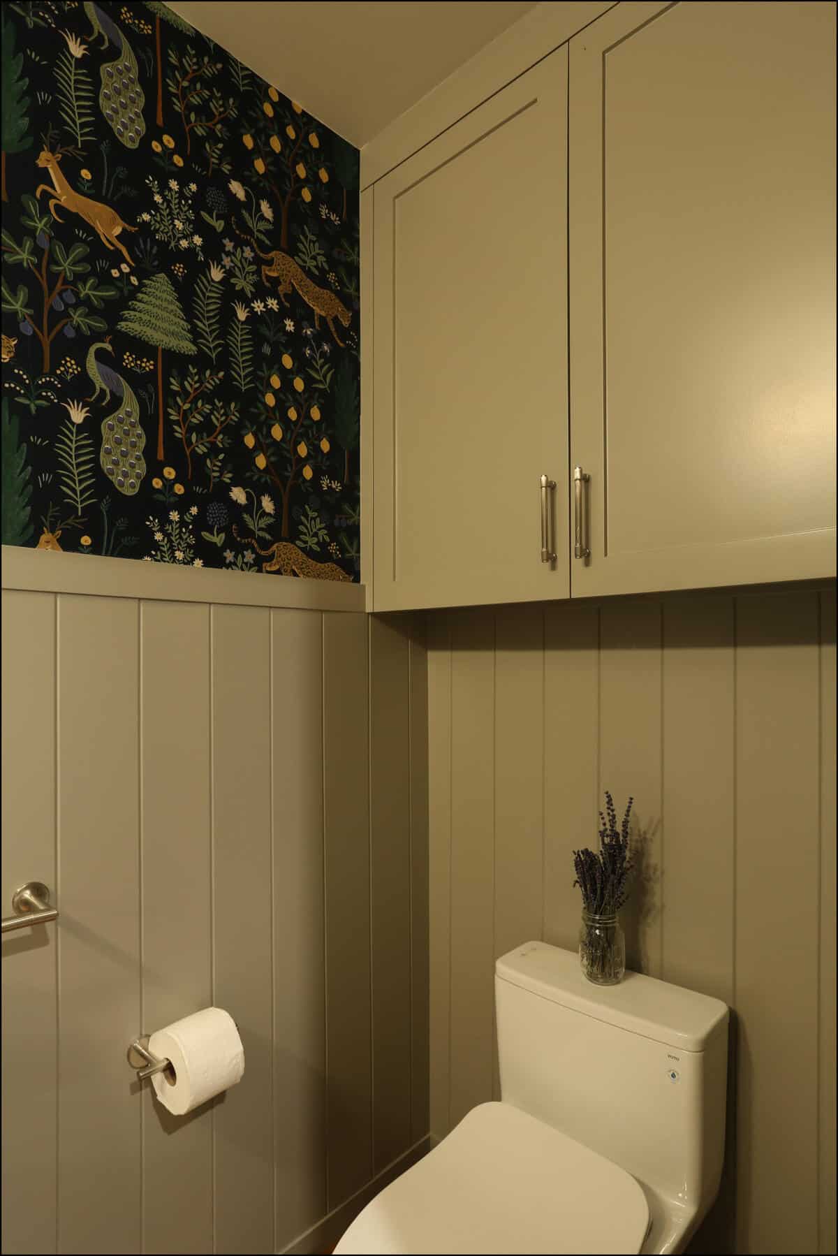 A small bathroom with taupe paneling, a white toilet, floral and animal-patterned wallpaper, cabinetry above, and a glass jar with lavender on the tank. Modern bathroom with a white toilet, gray paneling, and a vibrant patterned wallpaper featuring trees and animals, showcasing ReCraft Home Remodeling's design capabilities in whole house renovations.