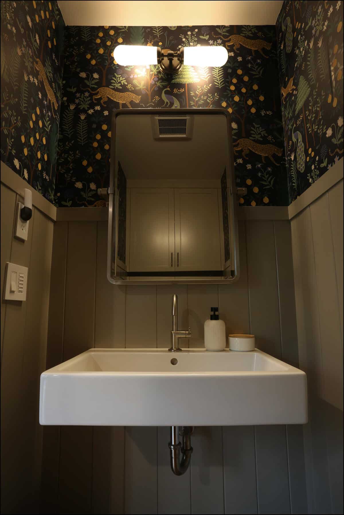 Bathroom with a modern white sink, mirror, and upper cabinet. Walls feature dark floral and animal print wallpaper with two overhead lights. Modern bathroom design featuring a sleek white sink, wall-mounted mirror, and decorative wallpaper with colorful patterns, showcasing ReCraft Home Remodeling's interior design capabilities in Portland, Oregon.