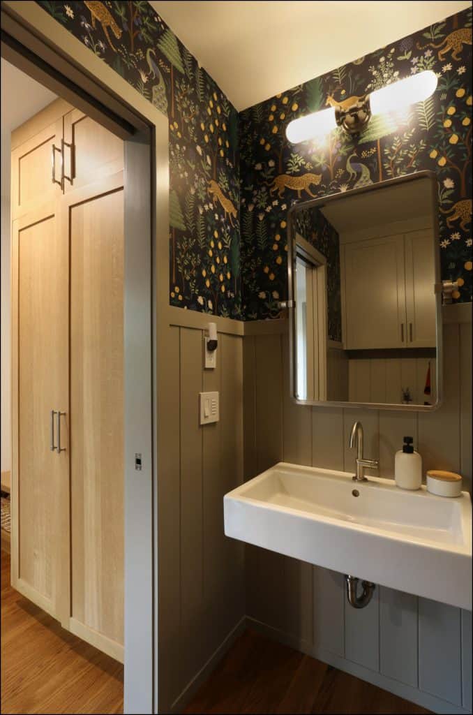Bathroom with a wall-mounted sink, patterned wallpaper, wall light above a rectangular mirror, and wooden accent panels. A view of a closet with wooden doors is visible through the open entryway. Bathroom with a wall-mounted sink, patterned wallpaper, wall light above a rectangular mirror, and wooden accent panels. A view of a closet with wooden doors is visible through the open entryway.