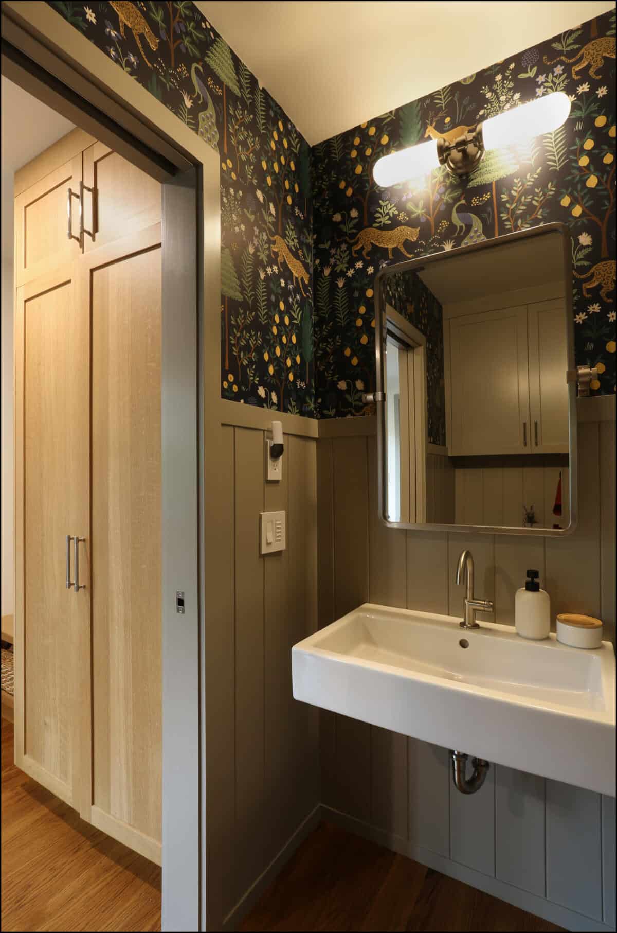 Bathroom with a wall-mounted sink, patterned wallpaper, wall light above a rectangular mirror, and wooden accent panels. A view of a closet with wooden doors is visible through the open entryway. Modern bathroom design featuring a sleek white sink, decorative wallpaper with nature motifs, wooden cabinetry, and a large mirror, showcasing ReCraft Home Remodeling's style in whole house renovations in Portland, Oregon.