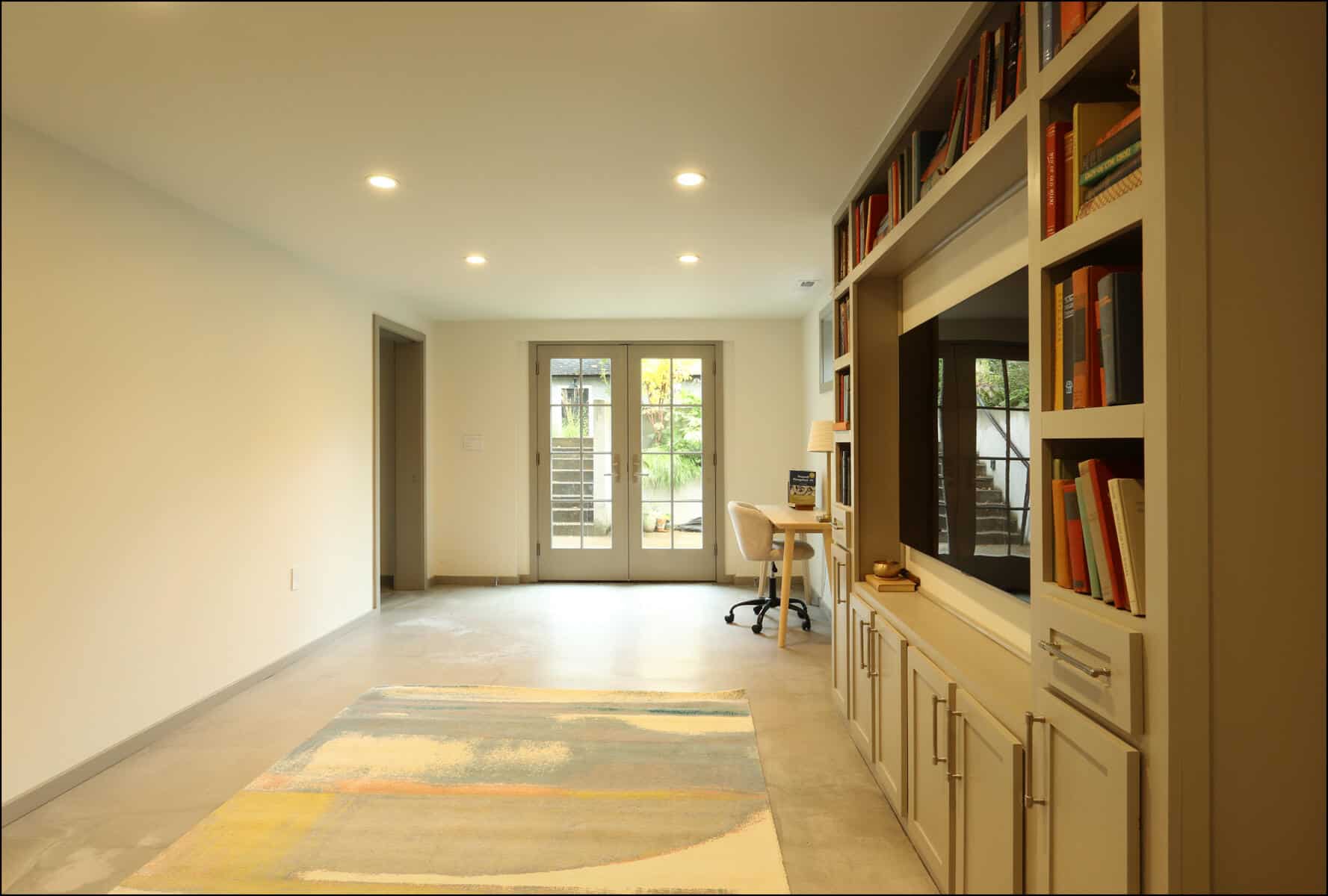 A minimalist room with built-in bookshelves, a desk, and a chair. Double doors lead outside. The floor is tiled with a rug. Modern interior of a remodeled room featuring a built-in bookshelf, desk area, and large windows, showcasing ReCraft Home Remodeling's design capabilities in Portland, Oregon.