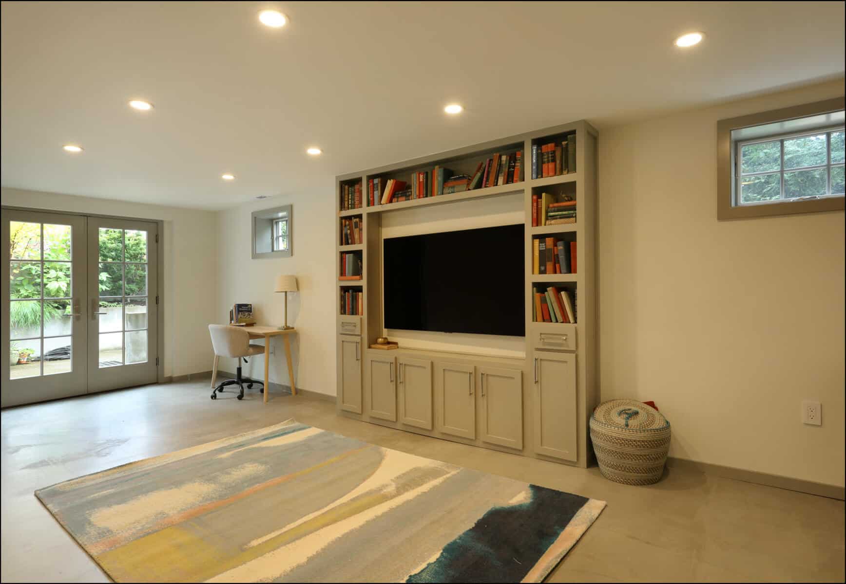 A modern living room features a wall-mounted TV, built-in bookshelves, a small desk and chair, a colorful rug, and glass doors leading outside. Modern living room with built-in shelves, a large TV, a work desk, and a colorful area rug, featuring natural light from windows and a door leading outside, showcasing ReCraft Home Remodeling's whole house renovation in Portland, OR.