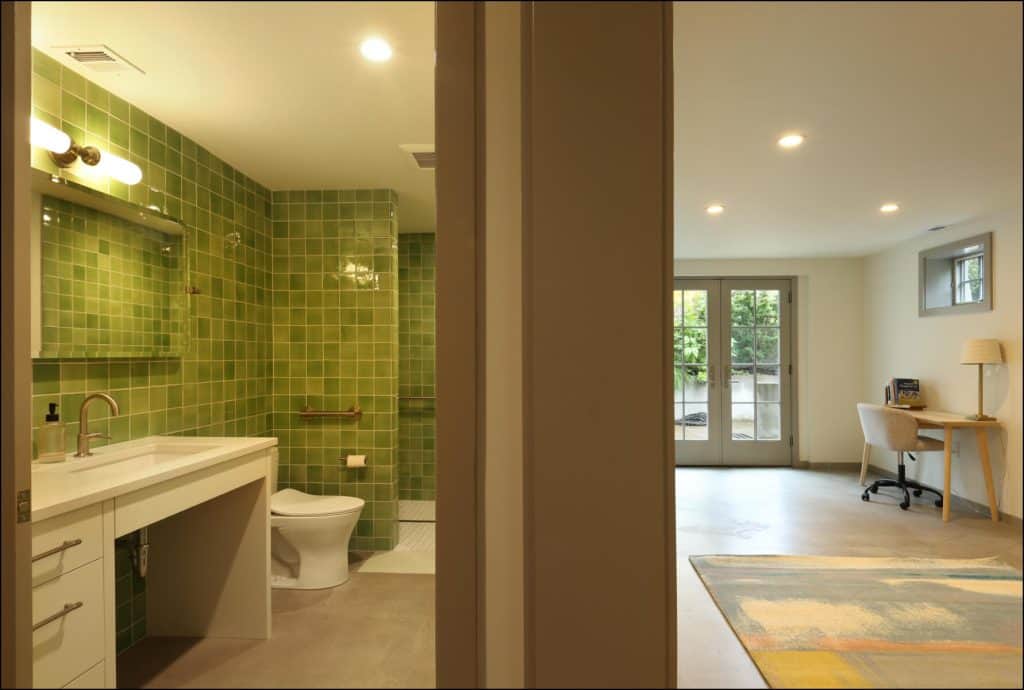 View of a bathroom with green tiled walls, toilet, and sink on the left, and a spacious room with a desk and chair on the right. View of a bathroom with green tiled walls, toilet, and sink on the left, and a spacious room with a desk and chair on the right.
