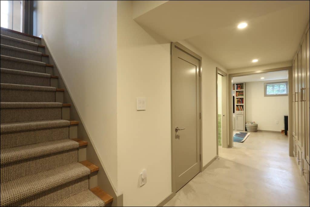 A basement hallway with carpeted stairs on the left, a closed door, and a view into a room with shelves and a small window. A basement hallway with carpeted stairs on the left, a closed door, and a view into a room with shelves and a small window.