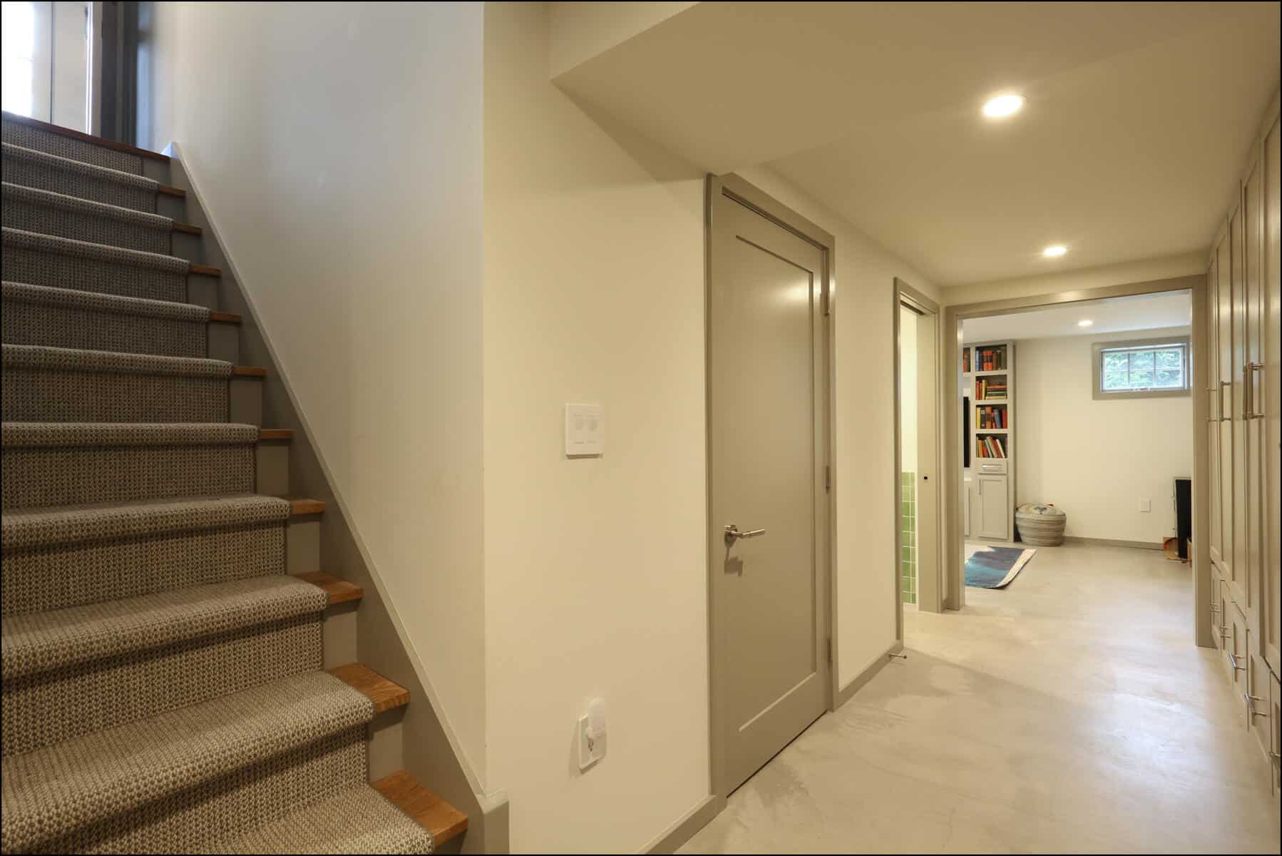 A basement hallway with carpeted stairs on the left, a closed door, and a view into a room with shelves and a small window. Modern interior hallway with staircase leading to an upper level, showcasing sleek design and ample lighting in a whole house remodel by ReCraft Home Remodeling in Portland, Oregon.
