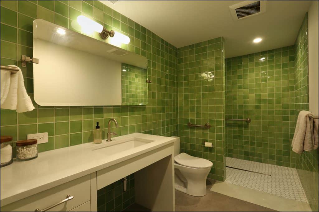 A bathroom with green tiled walls features a large mirror, a sink with a white countertop, a toilet, and a walk-in shower with a sliding glass door and a towel rack. A bathroom with green tiled walls features a large mirror, a sink with a white countertop, a toilet, and a walk-in shower with a sliding glass door and a towel rack.