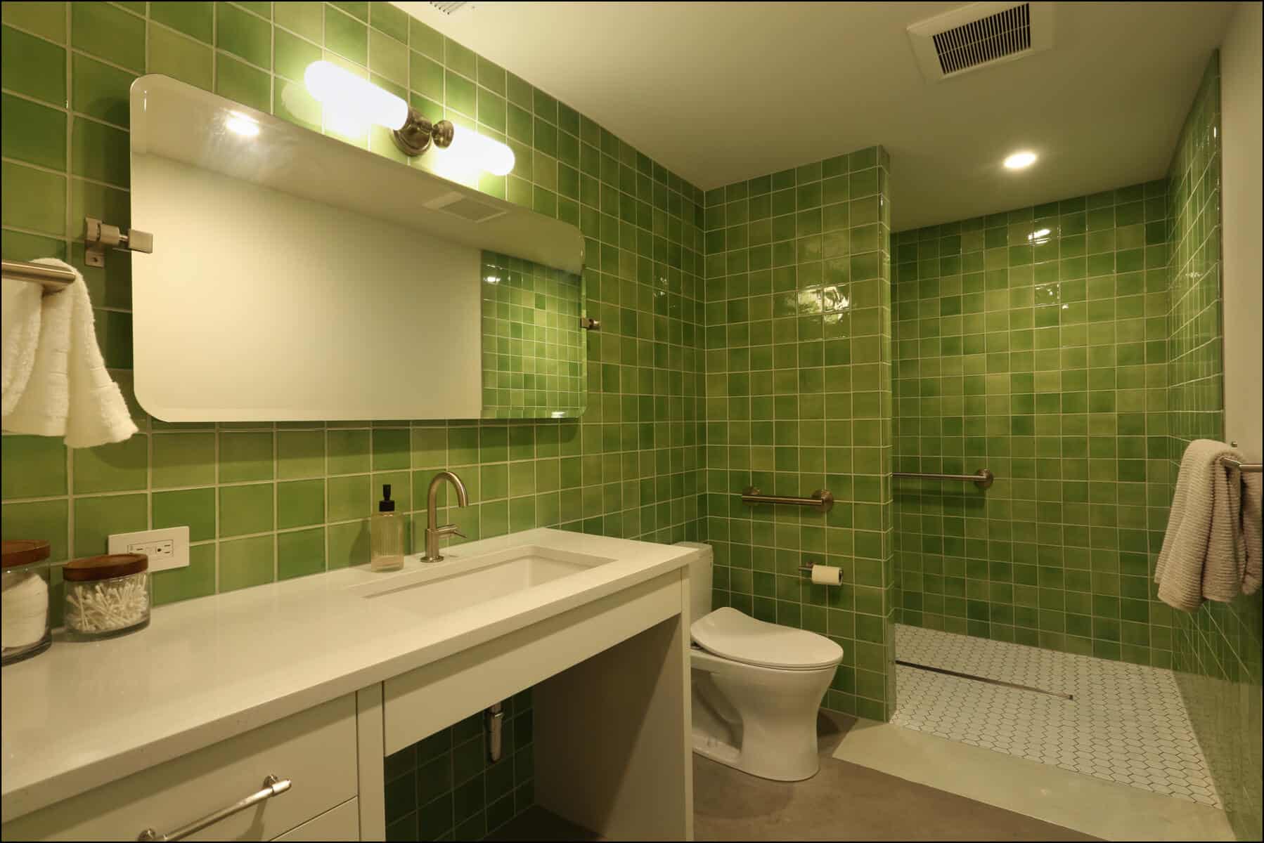 A bathroom with green tiled walls features a large mirror, a sink with a white countertop, a toilet, and a walk-in shower with a sliding glass door and a towel rack. Modern bathroom design featuring vibrant green tile walls, a sleek vanity with a mirror, and a spacious shower area, showcasing ReCraft Home Remodeling's expertise in whole house renovations.