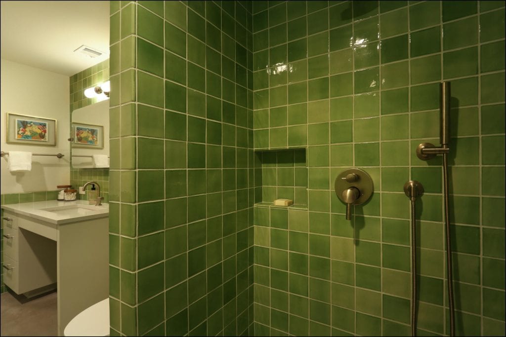 Bathroom with green tiled shower, bronze fixtures, and a soap niche; vanity with sink, mirror, and wall lights visible in the background. Bathroom with green tiled shower, bronze fixtures, and a soap niche; vanity with sink, mirror, and wall lights visible in the background.