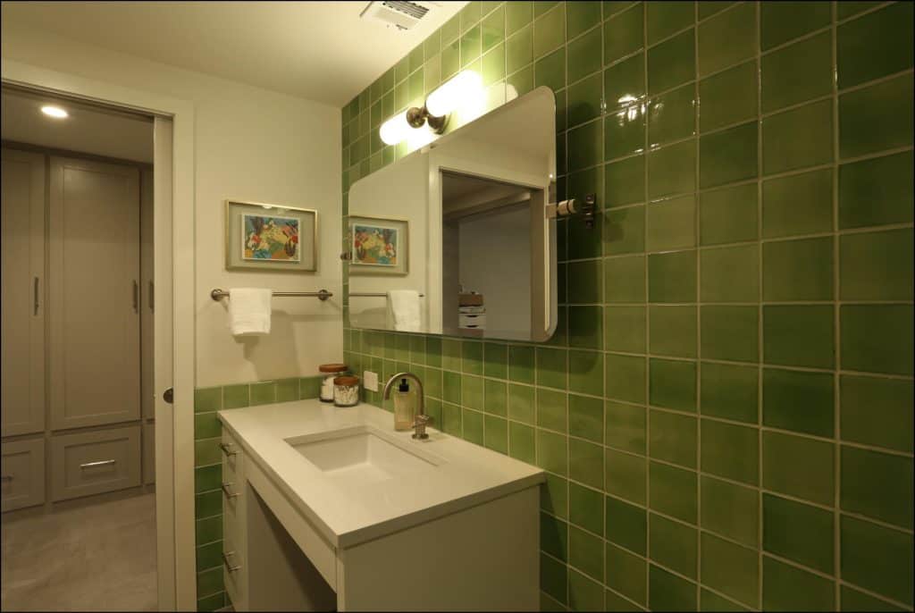 Bathroom with green tiled wall, white sink with silver faucet, and rectangular mirror. Two framed pictures and towel hang above the towel rack. Doorway leads to a closet area. Bathroom with green tiled wall, white sink with silver faucet, and rectangular mirror. Two framed pictures and towel hang above the towel rack. Doorway leads to a closet area.