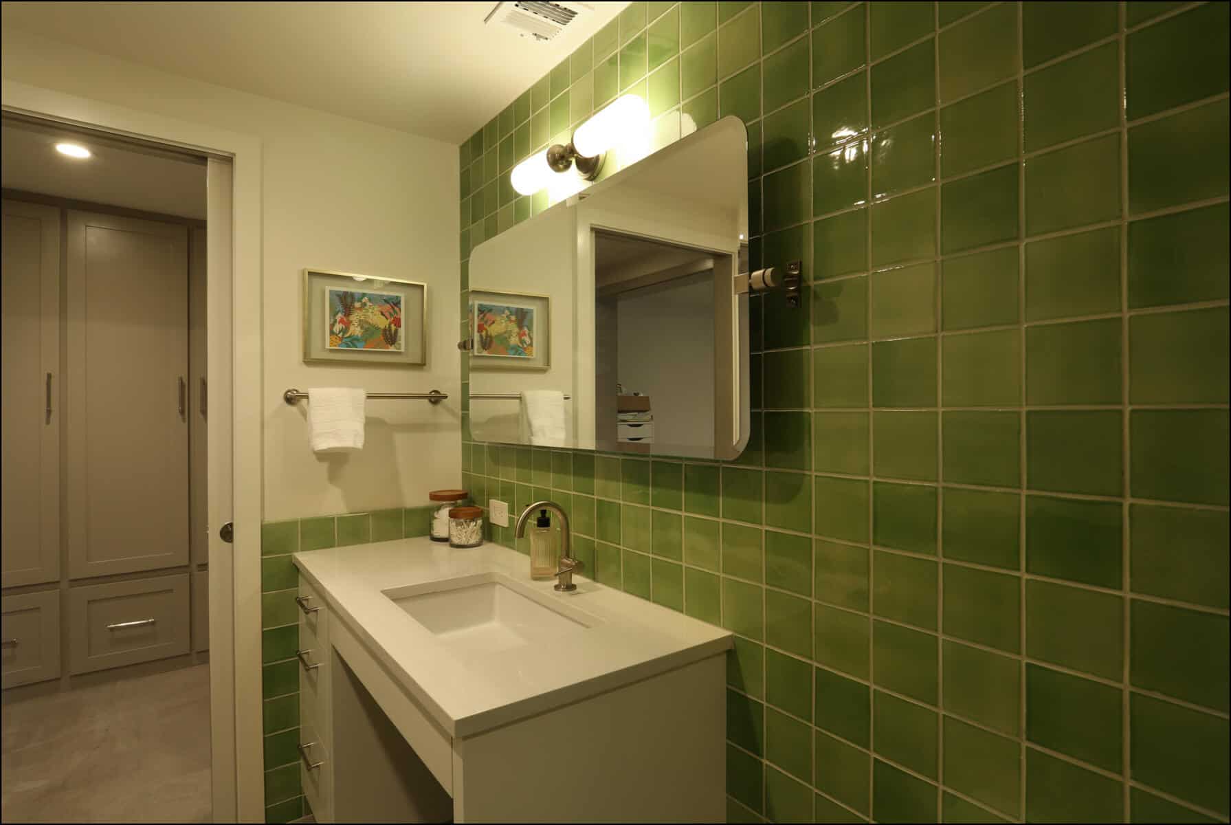 Bathroom with green tiled wall, white sink with silver faucet, and rectangular mirror. Two framed pictures and towel hang above the towel rack. Doorway leads to a closet area. Modern bathroom with green tiled walls, white vanity, mirror, and decorative artwork, showcasing ReCraft Home Remodeling's design aesthetic in Portland.