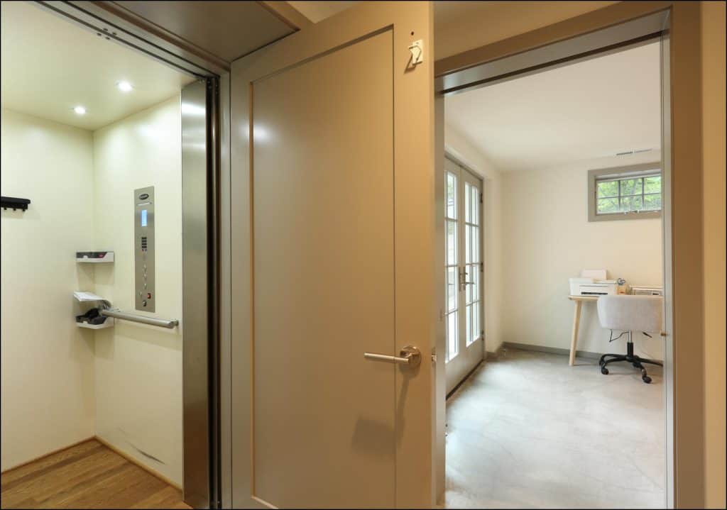 View of an open residential elevator next to a room with a desk and chair, illuminated by natural light from a window. View of an open residential elevator next to a room with a desk and chair, illuminated by natural light from a window.