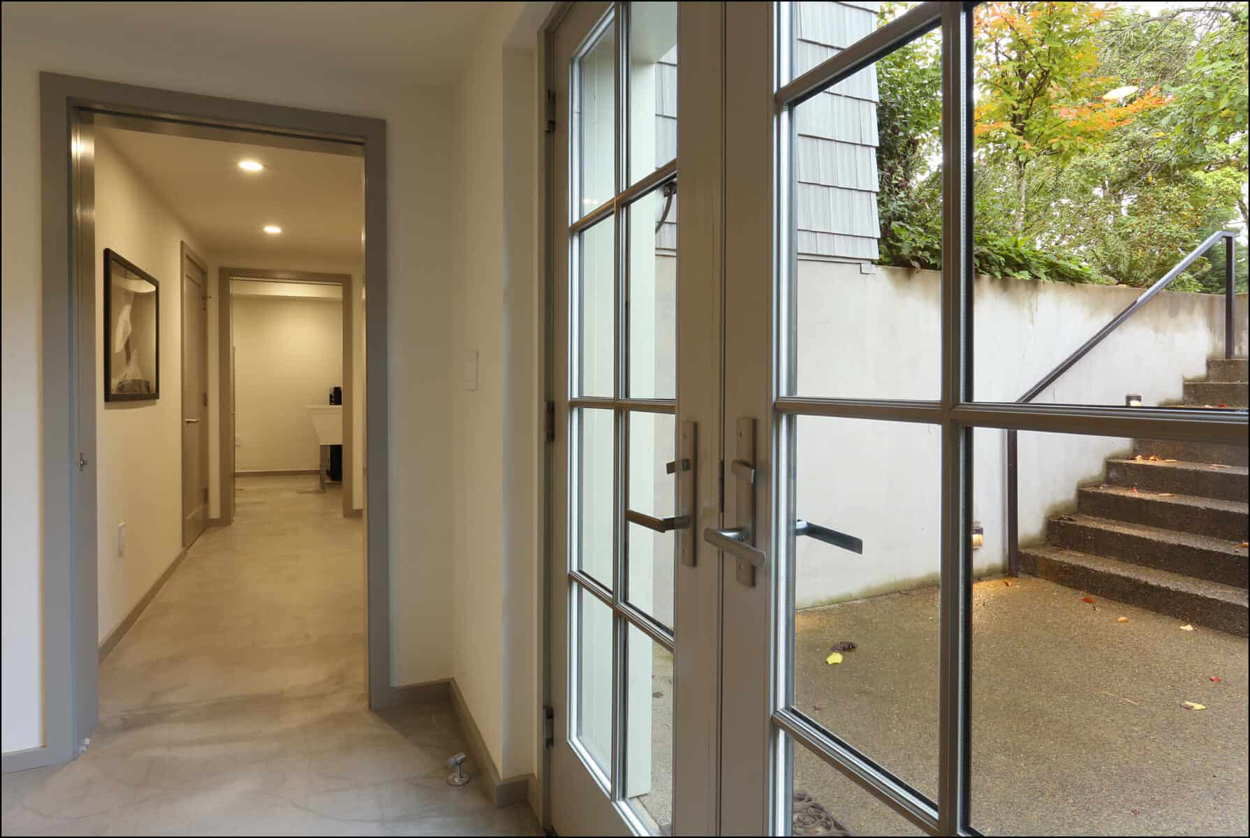 A hallway with tiled floor, open glass door leading outside to steps, and a framed picture on the wall. Entrance view of a modern home interior leading to outdoor stairs, showcasing a bright and inviting space, part of ReCraft Home Remodeling's whole house remodel projects in Portland, Oregon.