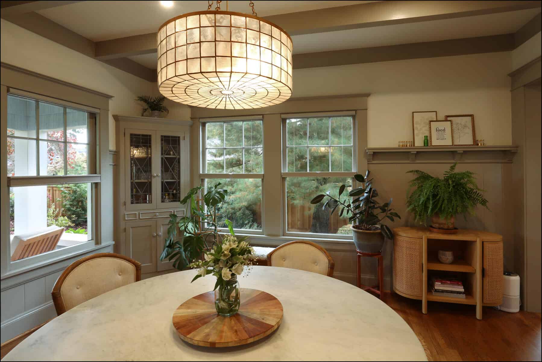 Cozy dining room with a round table, a vase of flowers, chairs, large windows, various plants, decorative items on shelves, and a large overhead light fixture. Cozy dining room with a round table, a vase of flowers, chairs, large windows, various plants, decorative items on shelves, and a large overhead light fixture.