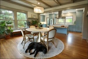 A cozy kitchen with light blue cabinets, a round dining table, and chairs on a rug. A black dog rests on the floor. Large windows and plants create a pleasant atmosphere.
