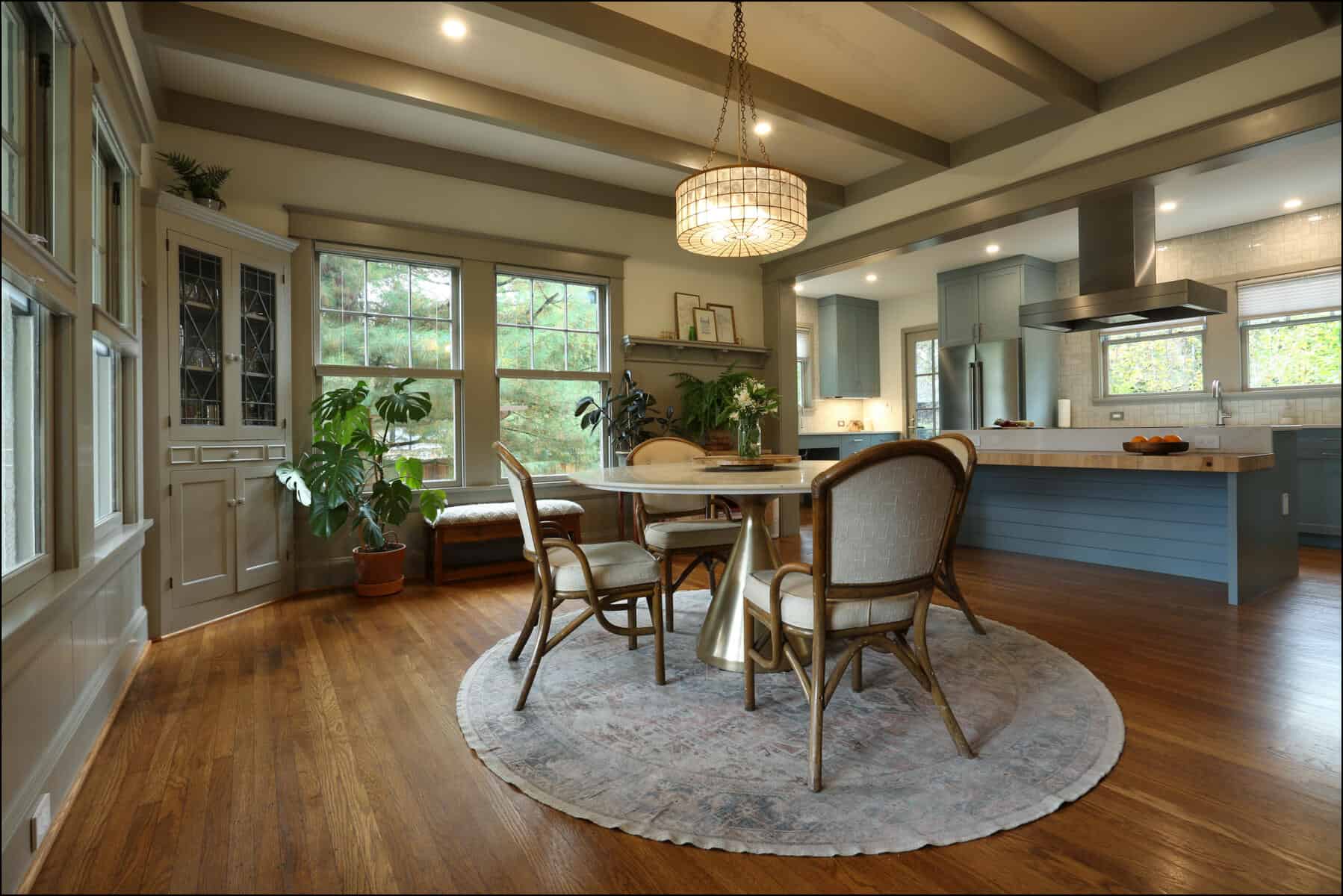 A modern kitchen with wooden floors, a round rug, and a dining set. Large windows, plants, and a chandelier enhance the decor. Cabinets and a kitchen island are visible in the background. A modern kitchen with wooden floors, a round rug, and a dining set. Large windows, plants, and a chandelier enhance the decor. Cabinets and a kitchen island are visible in the background.