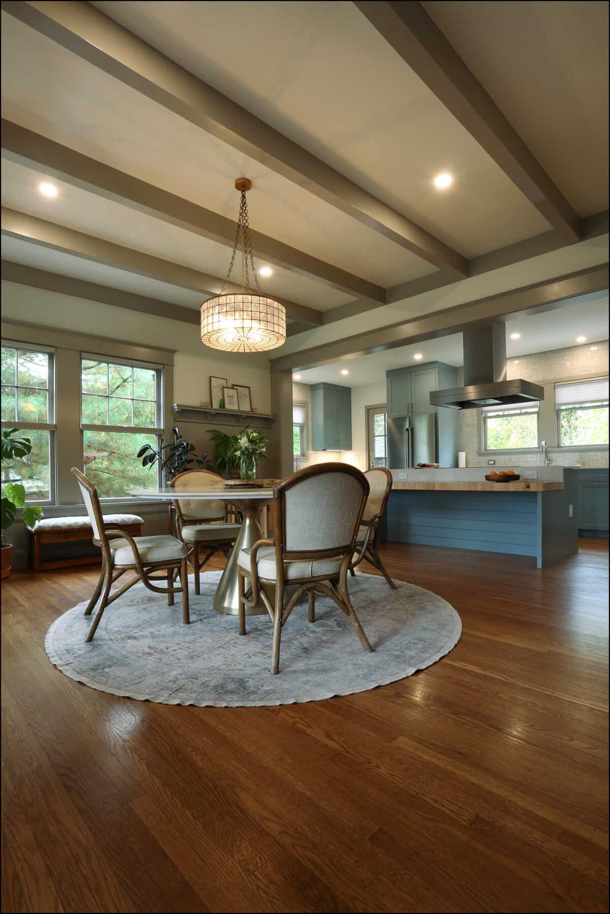A modern dining room with a round table and four chairs on a rug, wood flooring, and a chandelier. The kitchen with blue cabinets and island is visible in the background. A modern dining room with a round table and four chairs on a rug, wood flooring, and a chandelier. The kitchen with blue cabinets and island is visible in the background.