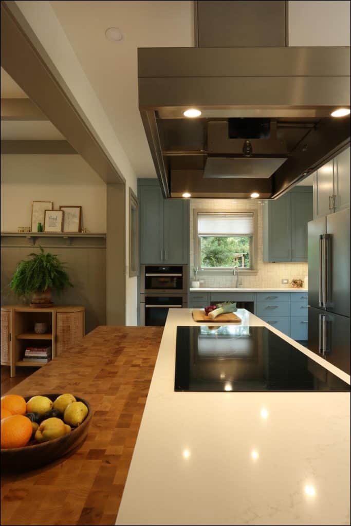 Modern kitchen with a large island featuring a cooktop and a wooden fruit bowl. Stainless steel appliances and light blue cabinets are visible, along with a window above the sink. Modern kitchen with a large island featuring a cooktop and a wooden fruit bowl. Stainless steel appliances and light blue cabinets are visible, along with a window above the sink.