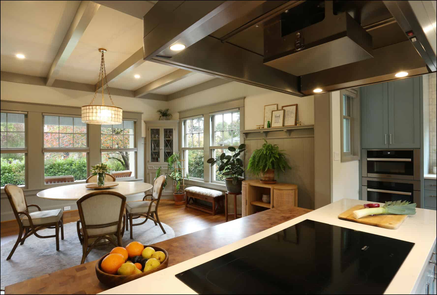 A modern kitchen with a wooden island, fruit bowl, and induction cooktop. In the background, a dining table with chairs is near large windows and indoor plants. A modern kitchen with a wooden island, fruit bowl, and induction cooktop. In the background, a dining table with chairs is near large windows and indoor plants.