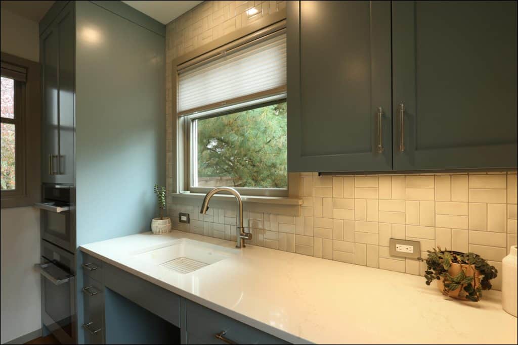 Modern kitchen with green cabinets, white countertop, and tile backsplash. A window with a blind above the sink, a small plant, and a potted vine on the counter. Modern kitchen with green cabinets, white countertop, and tile backsplash. A window with a blind above the sink, a small plant, and a potted vine on the counter.