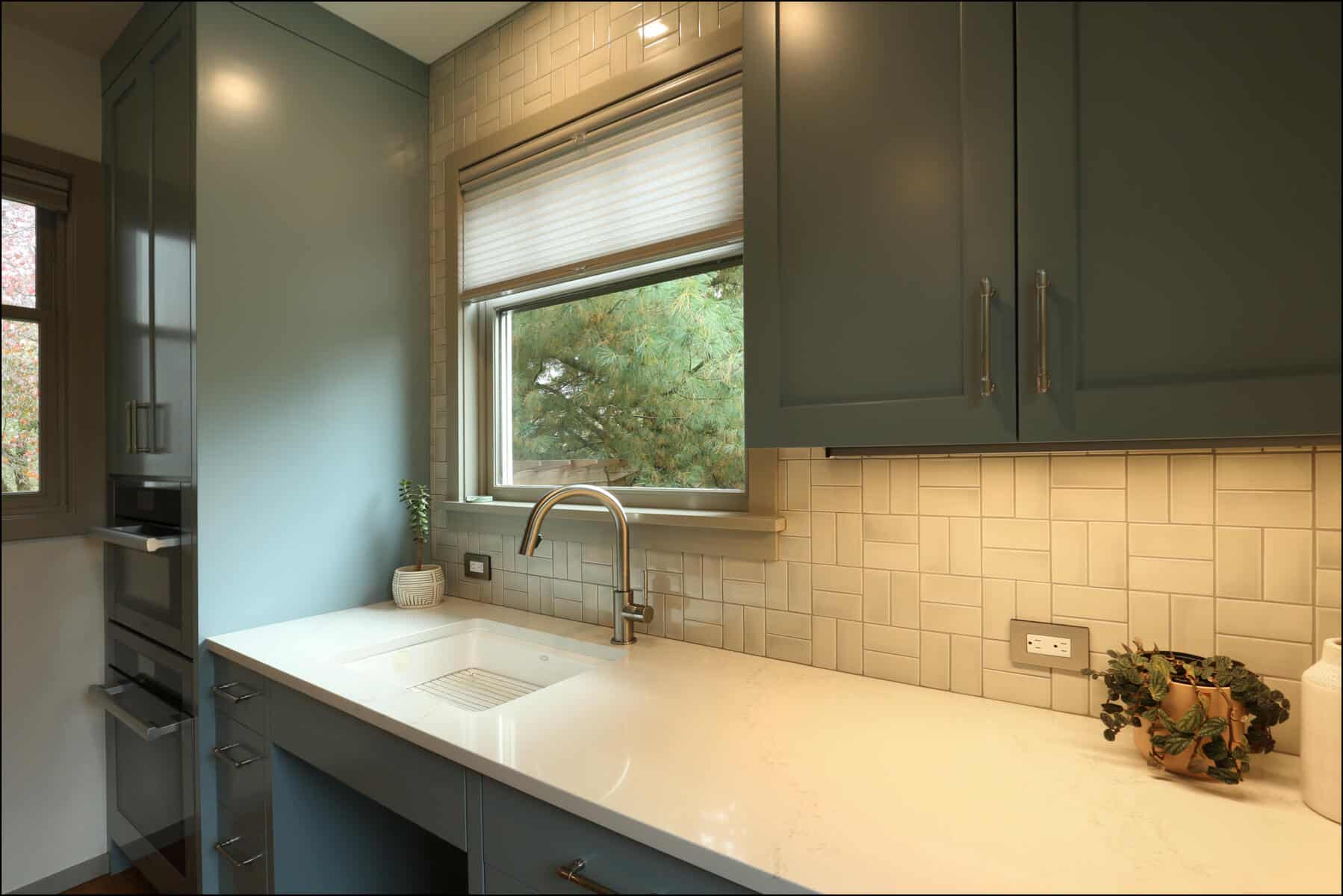 Modern kitchen with green cabinets, white countertop, and tile backsplash. A window with a blind above the sink, a small plant, and a potted vine on the counter. Modern kitchen with green cabinets, white countertop, and tile backsplash. A window with a blind above the sink, a small plant, and a potted vine on the counter.