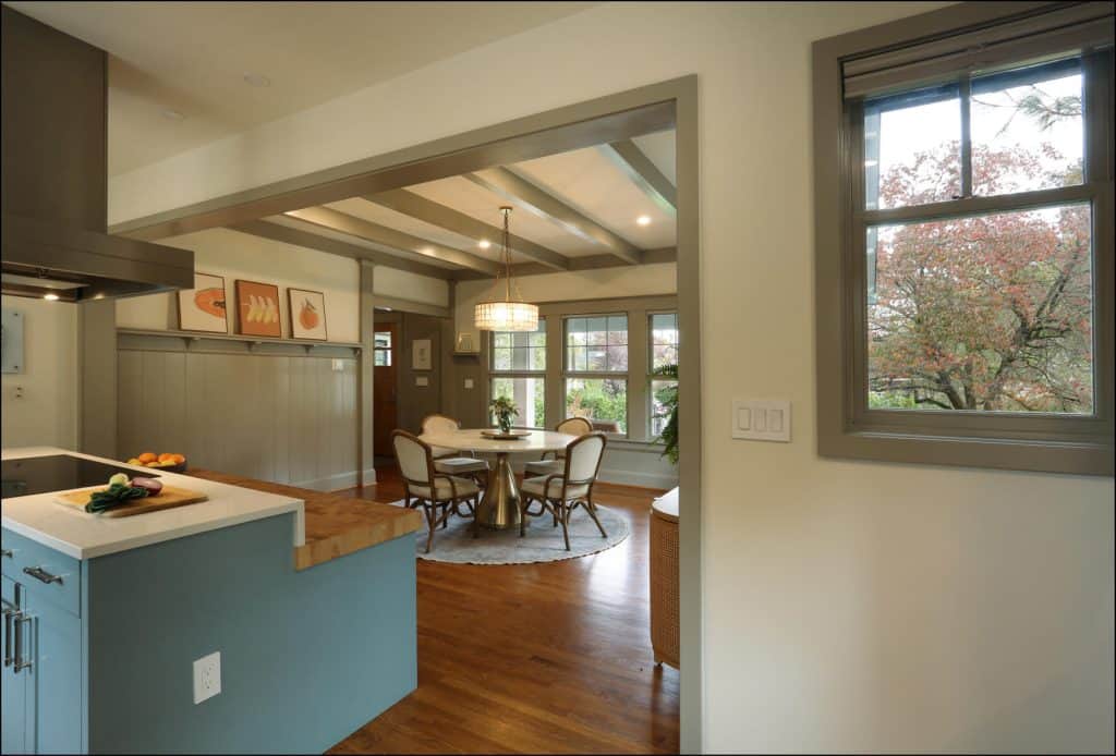 Open kitchen view leading to a dining area with a round table, surrounded by chairs. Large windows offer a view of trees outside. Walls feature framed artwork. Wood floors throughout. Open kitchen view leading to a dining area with a round table, surrounded by chairs. Large windows offer a view of trees outside. Walls feature framed artwork. Wood floors throughout.