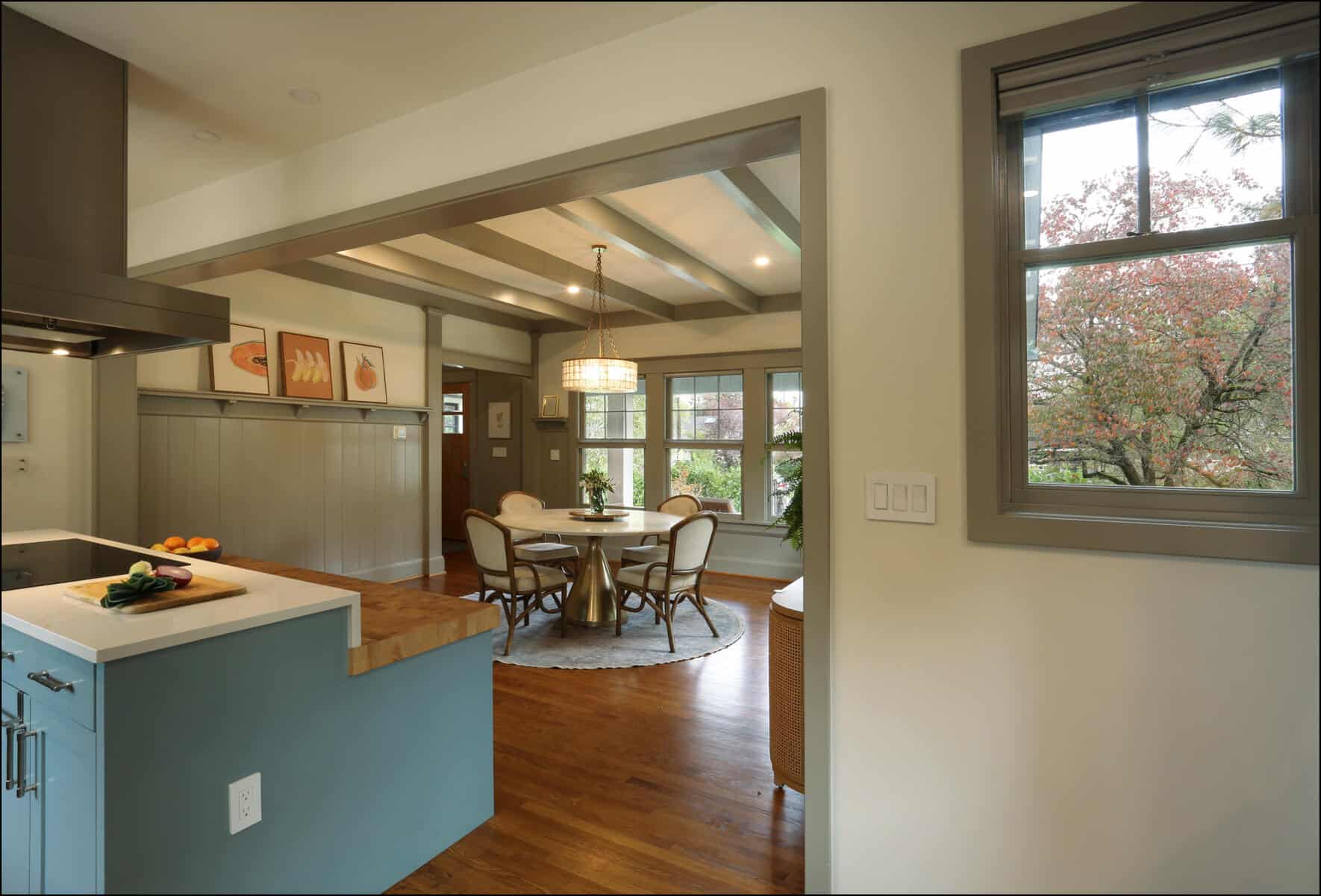 Open kitchen view leading to a dining area with a round table, surrounded by chairs. Large windows offer a view of trees outside. Walls feature framed artwork. Wood floors throughout. Open kitchen view leading to a dining area with a round table, surrounded by chairs. Large windows offer a view of trees outside. Walls feature framed artwork. Wood floors throughout.