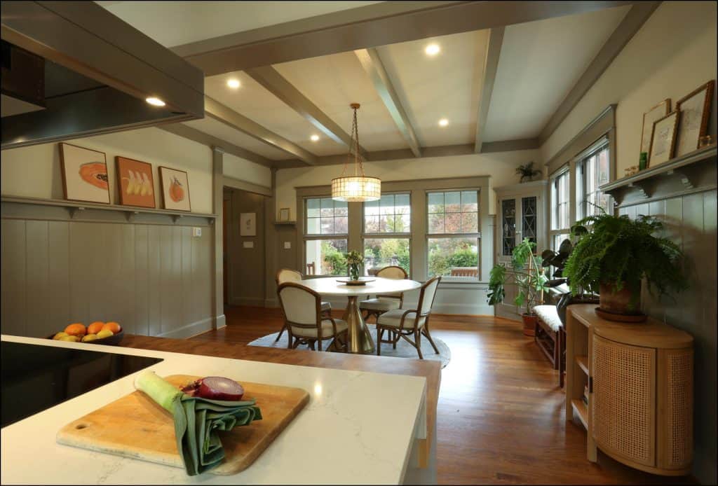 A spacious dining room with a table and chairs, large windows, plants, and wall art. A cutting board with vegetables is on the kitchen counter in the foreground. A spacious dining room with a table and chairs, large windows, plants, and wall art. A cutting board with vegetables is on the kitchen counter in the foreground.