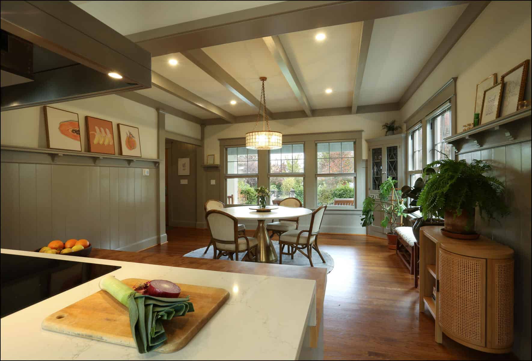 A spacious dining room with a table and chairs, large windows, plants, and wall art. A cutting board with vegetables is on the kitchen counter in the foreground. A spacious dining room with a table and chairs, large windows, plants, and wall art. A cutting board with vegetables is on the kitchen counter in the foreground.