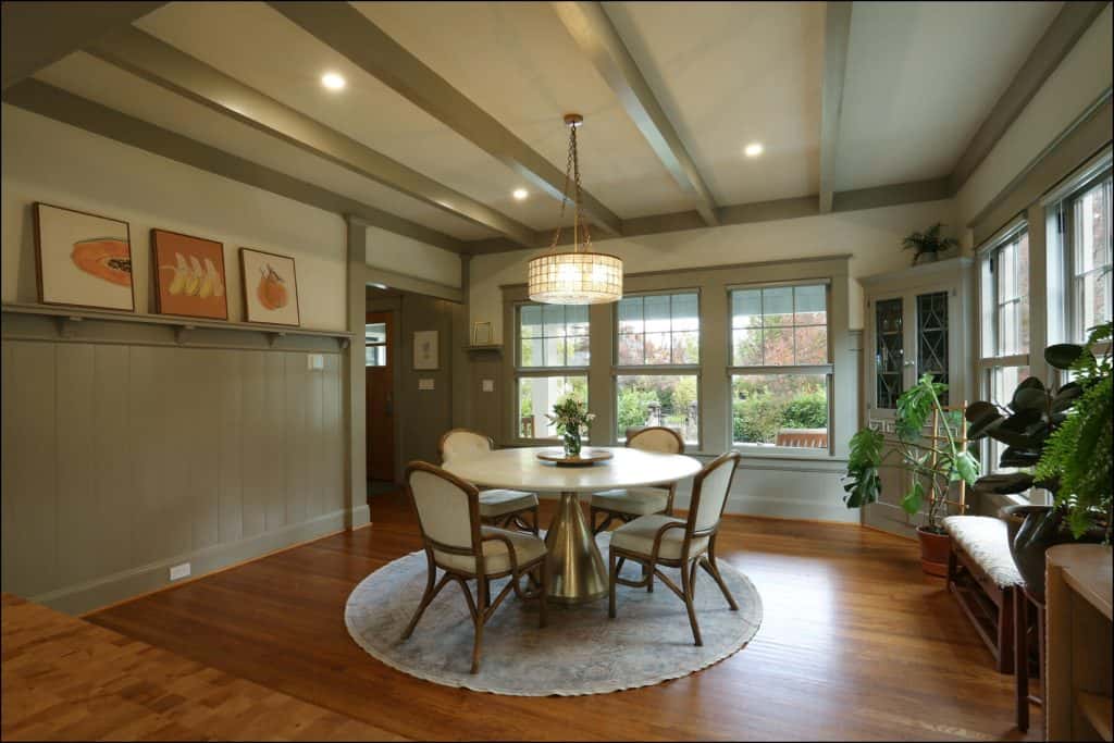 A dining room with a round table and four chairs on a rug. Large windows, a chandelier, potted plants, and wall paintings complete the decor. A dining room with a round table and four chairs on a rug. Large windows, a chandelier, potted plants, and wall paintings complete the decor.