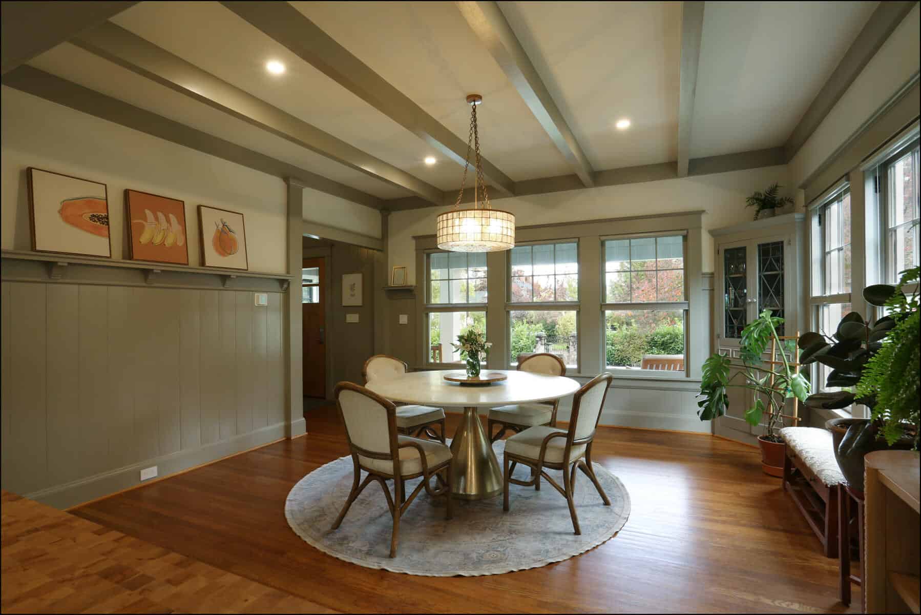 A dining room with a round table and four chairs on a rug. Large windows, a chandelier, potted plants, and wall paintings complete the decor. A dining room with a round table and four chairs on a rug. Large windows, a chandelier, potted plants, and wall paintings complete the decor.