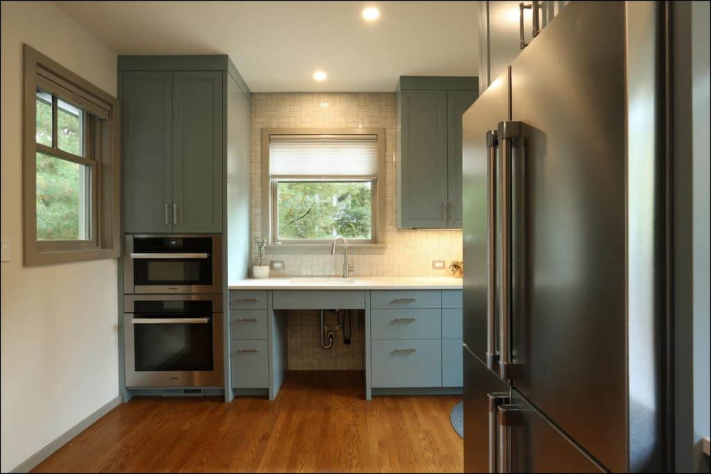 A modern kitchen with light gray cabinets, a large refrigerator, built-in oven, and hardwood floor. A window above the sink offers a view of greenery outside. A modern kitchen with light gray cabinets, a large refrigerator, built-in oven, and hardwood floor. A window above the sink offers a view of greenery outside.