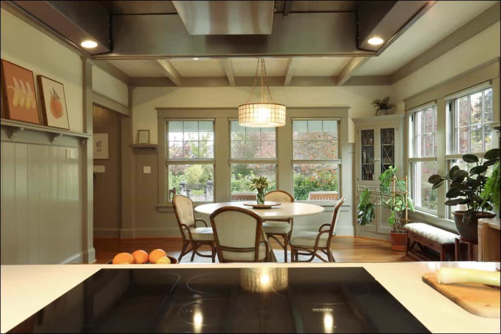 A cozy kitchen and dining area with a round table, chairs, and several plants. Large windows overlook a garden, and an induction cooktop is visible in the foreground. A cozy kitchen and dining area with a round table, chairs, and several plants. Large windows overlook a garden, and an induction cooktop is visible in the foreground.