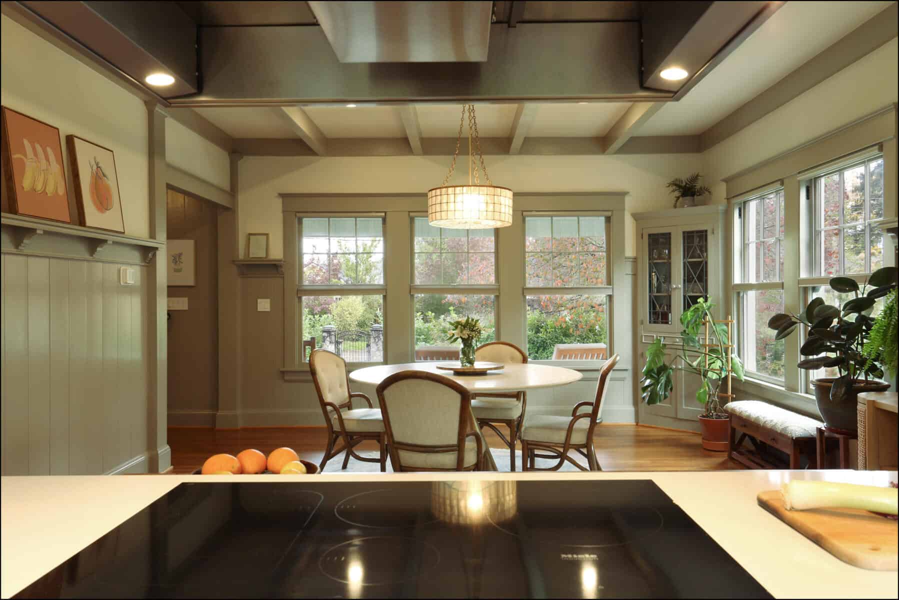 A cozy kitchen and dining area with a round table, chairs, and several plants. Large windows overlook a garden, and an induction cooktop is visible in the foreground. A cozy kitchen and dining area with a round table, chairs, and several plants. Large windows overlook a garden, and an induction cooktop is visible in the foreground.