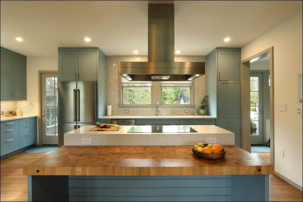 Modern kitchen with teal cabinets, a large island, wood countertops, a stainless steel refrigerator, and a range hood. A bowl of fruit and a cutting board are on the island. Modern kitchen with teal cabinets, a large island, wood countertops, a stainless steel refrigerator, and a range hood. A bowl of fruit and a cutting board are on the island.