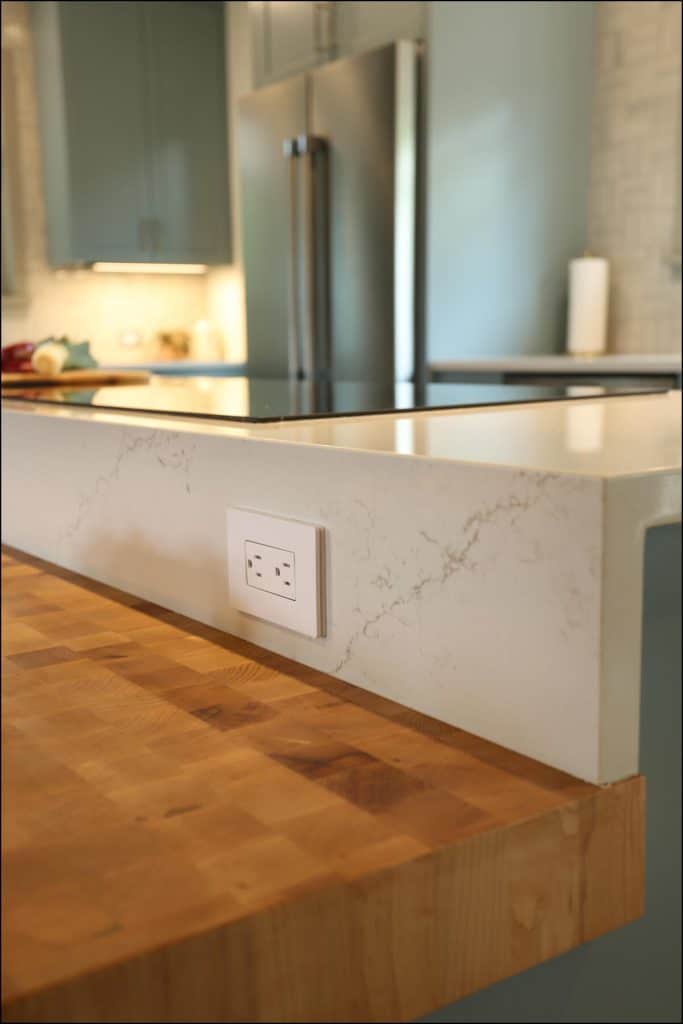 A kitchen counter with a marble finish and a wooden edge features a built-in electrical outlet. A stainless steel refrigerator is in the background. A kitchen counter with a marble finish and a wooden edge features a built-in electrical outlet. A stainless steel refrigerator is in the background.