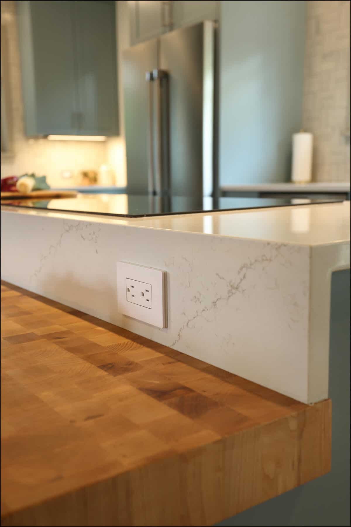 A kitchen counter with a marble finish and a wooden edge features a built-in electrical outlet. A stainless steel refrigerator is in the background. A kitchen counter with a marble finish and a wooden edge features a built-in electrical outlet. A stainless steel refrigerator is in the background.