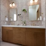 Modern bathroom with double sink vanity, brass hardware, rectangular mirrors, potted plants, and speckled wall tiles. Two wall sconces above mirrors provide lighting.