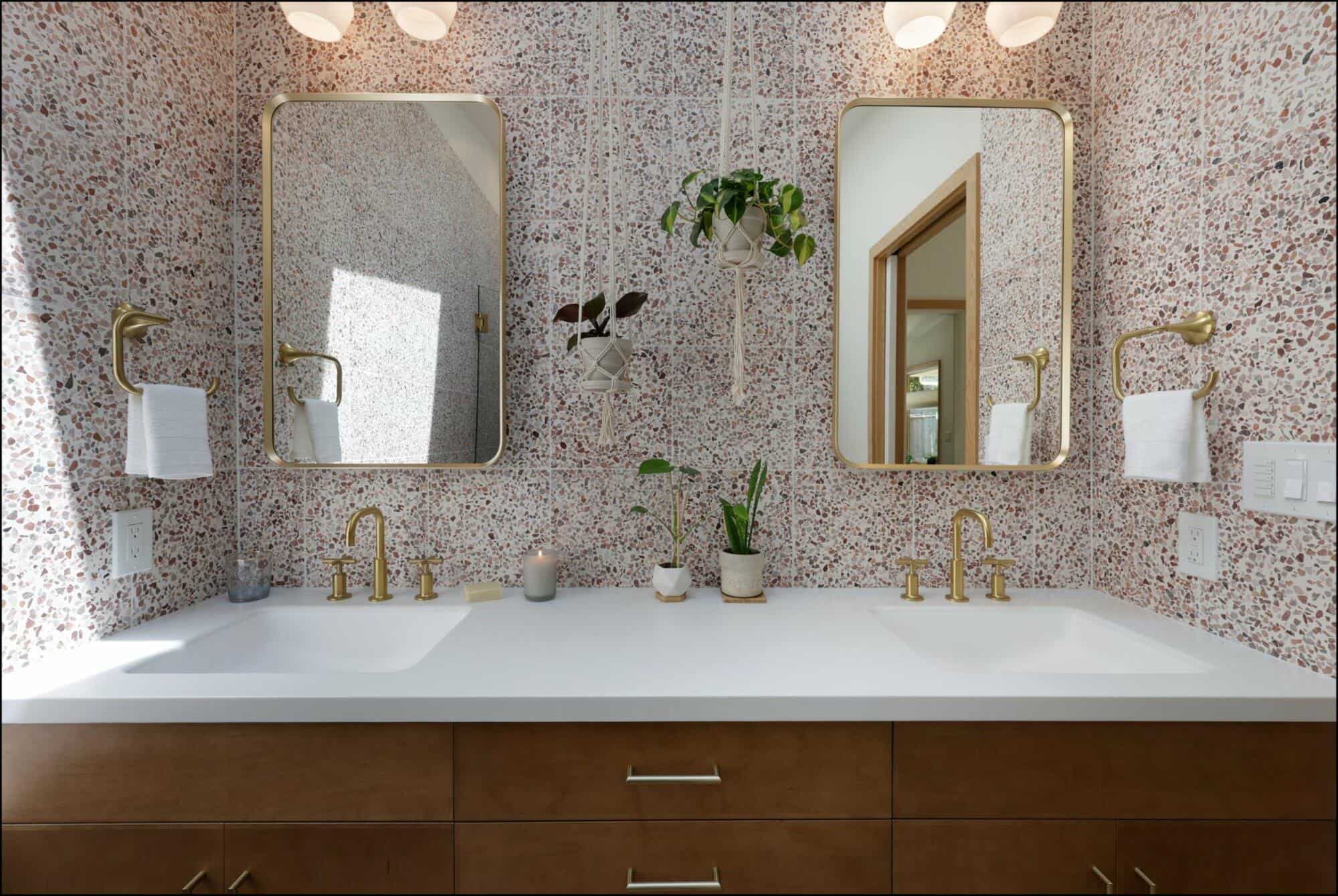 Double-sink bathroom vanity with brass fixtures, rectangular mirrors, a terrazzo backsplash, and hanging plants.